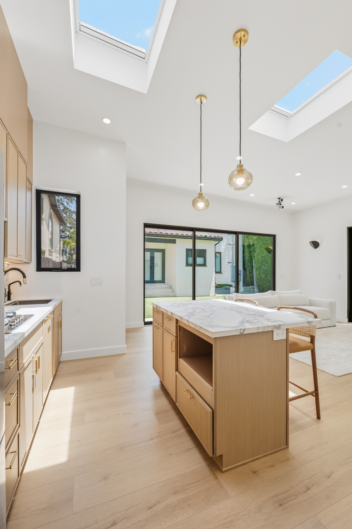- Oasis Builders, Inc. Bright open-concept kitchen with a marble island, light wood cabinets, and pendant lights, with large glass doors to the yard.