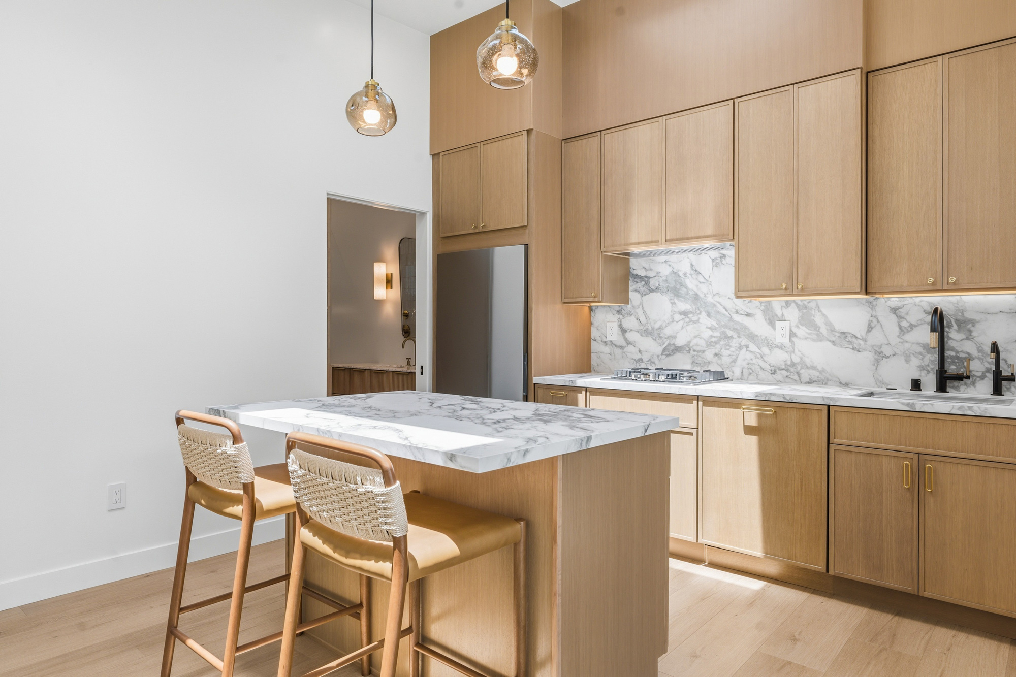 custom-oak-cabinetry-and-waterfall-island-adu-kitchen - Oasis Builders, Inc. Modern kitchen with light wood cabinets, marble countertops, an island with two wicker barstools and pendant lights hanging above.