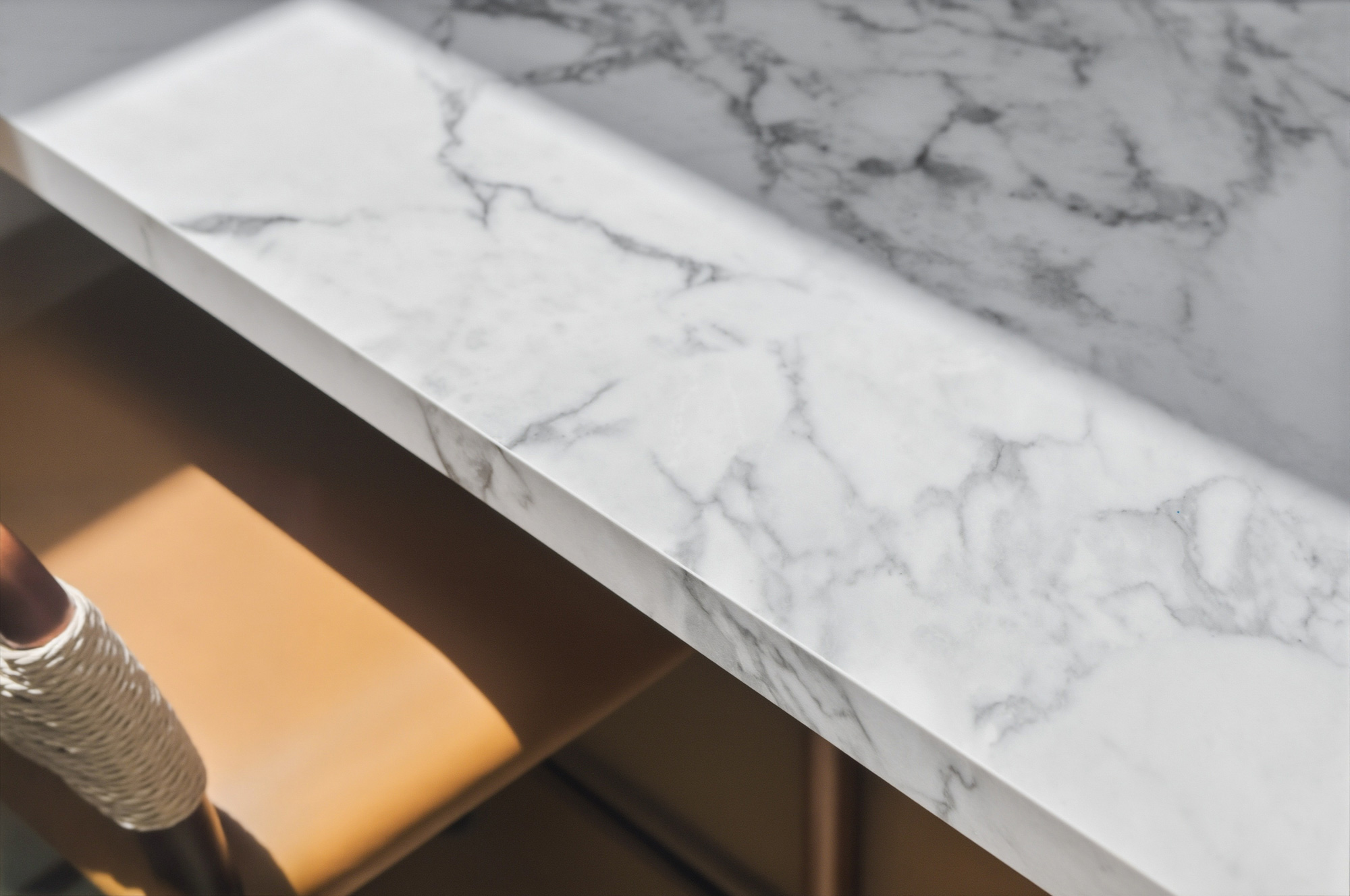 honed-porcelain-waterfall-island-countertop-detail - Oasis Builders, Inc. Close-up of a white marble countertop edge with gray veining, and a rope-wrapped support beneath in warm light.