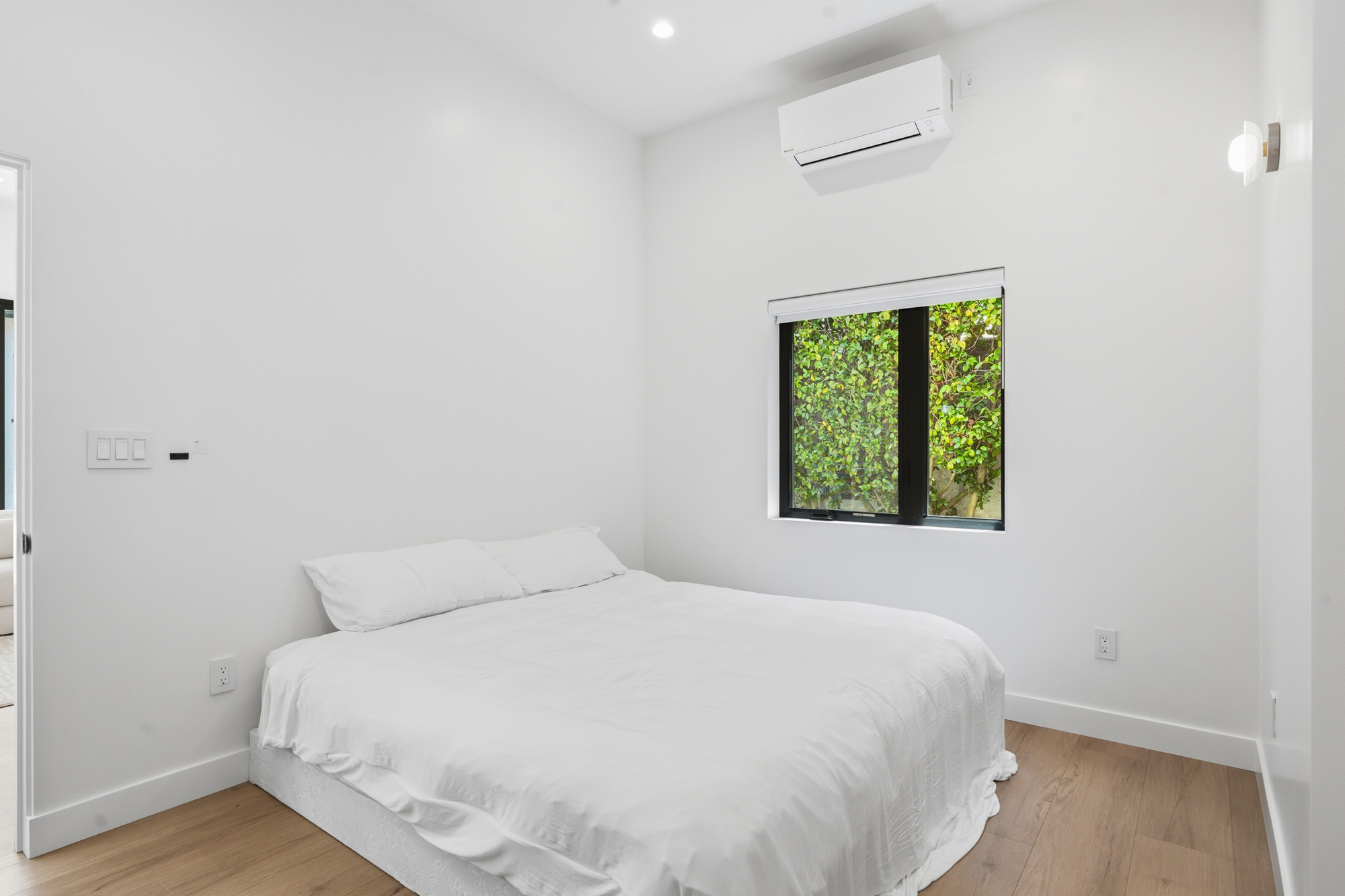 mid-city-los-angeles-adu-bedroom - Oasis Builders, Inc. Minimalist white bedroom with a neatly made bed, light wood floor, and a window showing green foliage outside