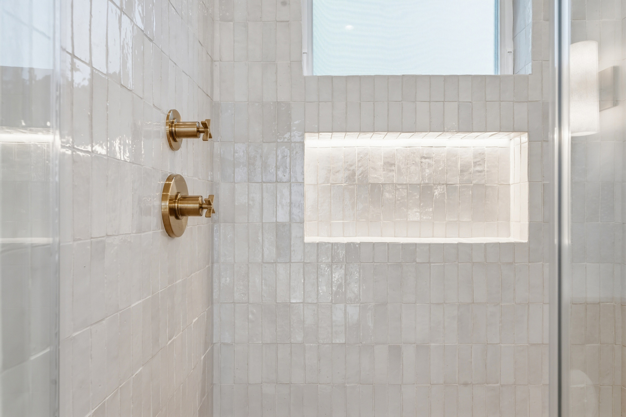 mid-city-los-angeles-led-shower-niche-detail - Oasis Builders, Inc. White-tiled shower with brass faucet handles on the left and a recessed shelf built into the wall, seen through a glass enclosure.