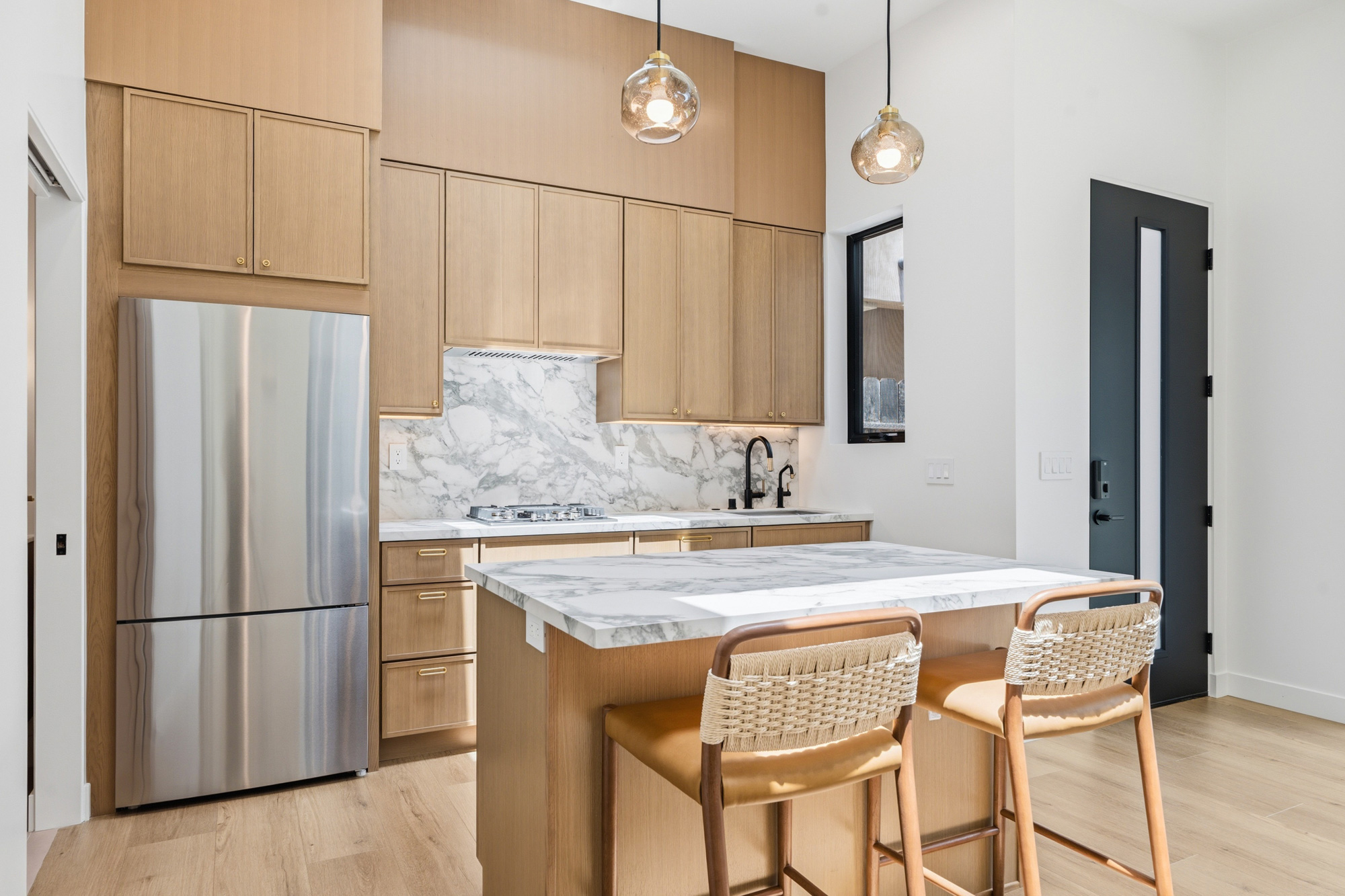 mid-city-los-angeles-oak-kitchen-cabinetry - Oasis Builders, Inc. Modern kitchen with tall wooden cabinets, marble countertops and backsplash, stainless steel refrigerator, and an island with two wicker bar stools.
