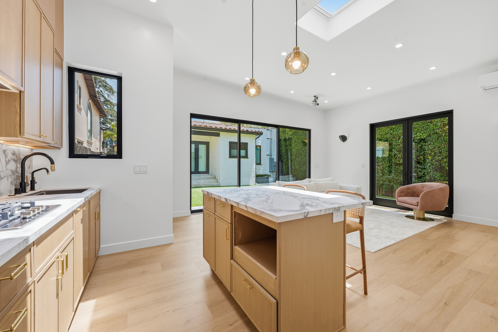 mid-city-los-angeles-scandinavian-adu-interior - Oasis Builders, Inc. Open-plan kitchen with a marble island, light wood cabinets, and large glass doors overlooking a garden/yard.