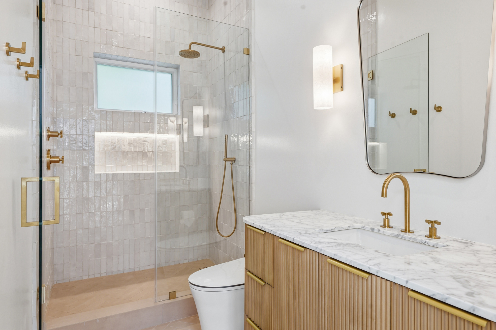 mid-city-los-angeles-zellige-tile-shower - Oasis Builders, Inc. Modern bathroom with a glass shower enclosure, white tiles, and brass fixtures alongside a marble countertop vanity.