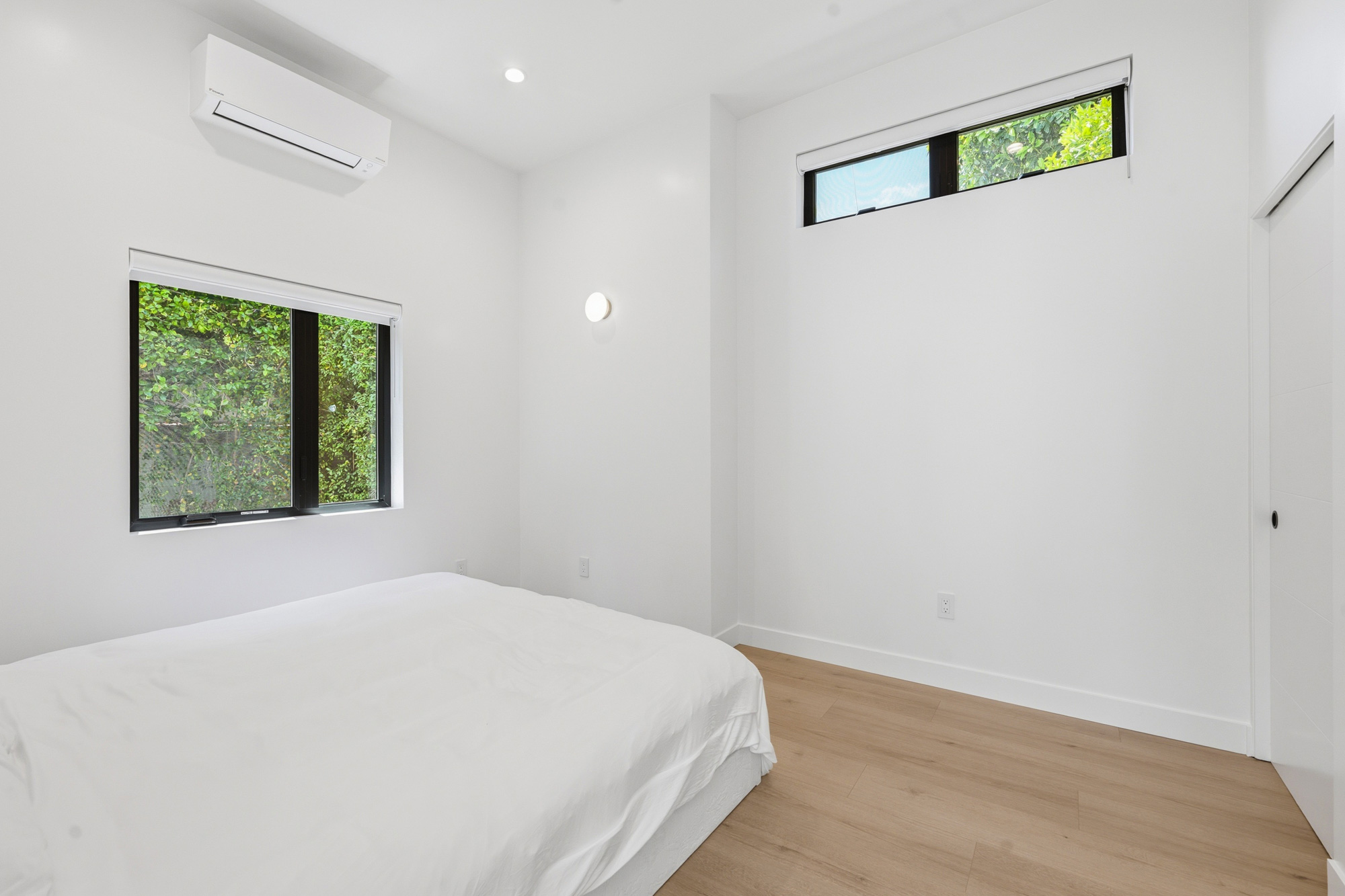 modern-adu-bedroom-with-high-ceilings-and-clerestory-window - Oasis Builders, Inc. Minimal white bedroom with a low white bed, wooden floor, and two rectangular windows showing greenery outside.