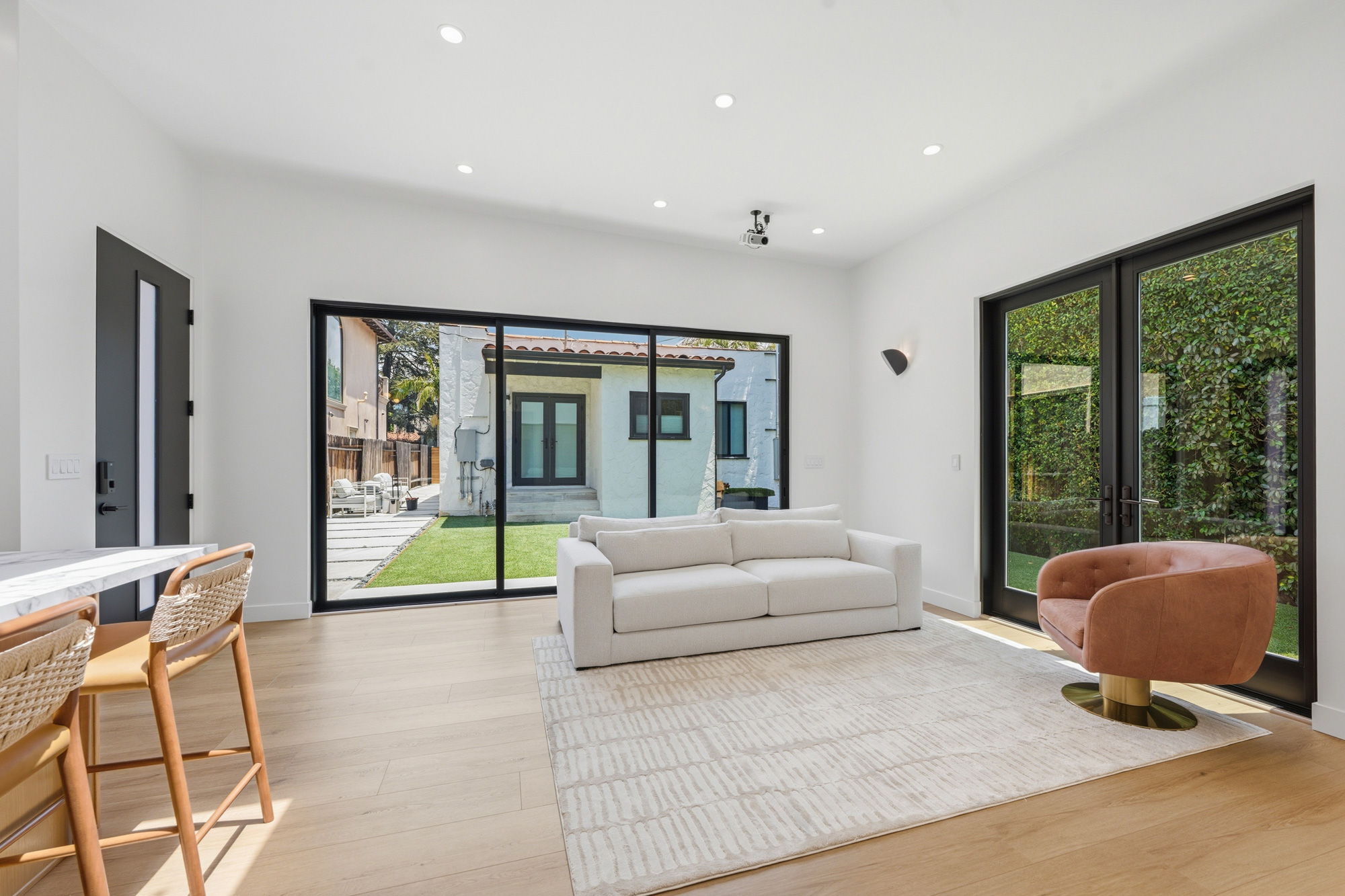 open-concept-adu-living-room-with-sliding-glass-doors - Oasis Builders, Inc. Bright contemporary living room with white walls, beige sofa, and large black-framed sliding glass doors to a green backyard.