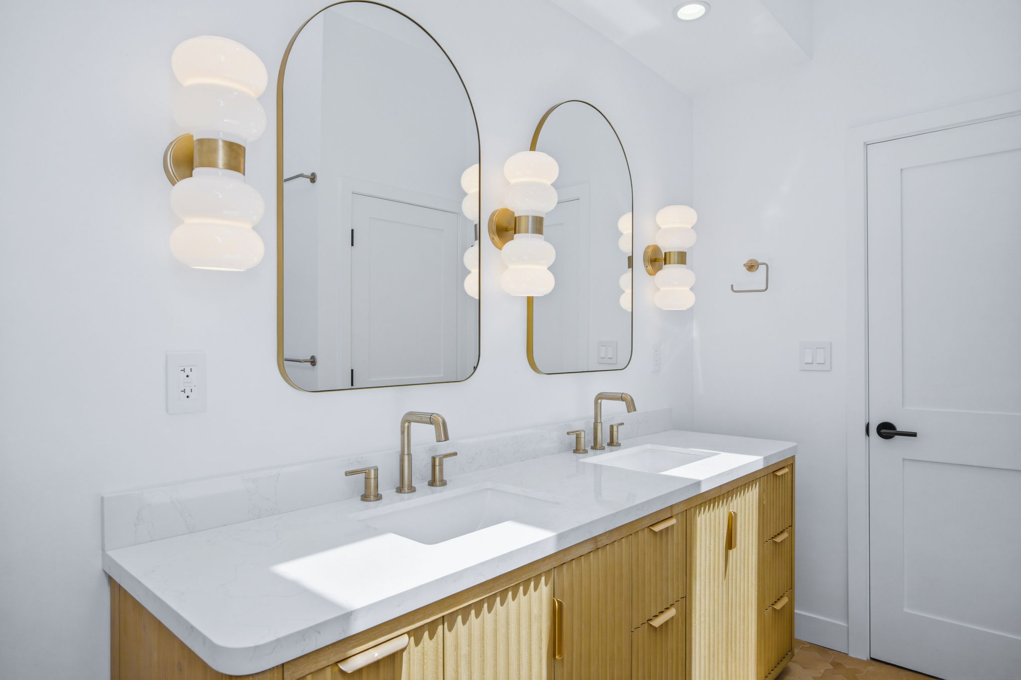 - Oasis Builders, Inc. Double vanity with white marble countertop, brass faucets, and light wood cabinets beneath.