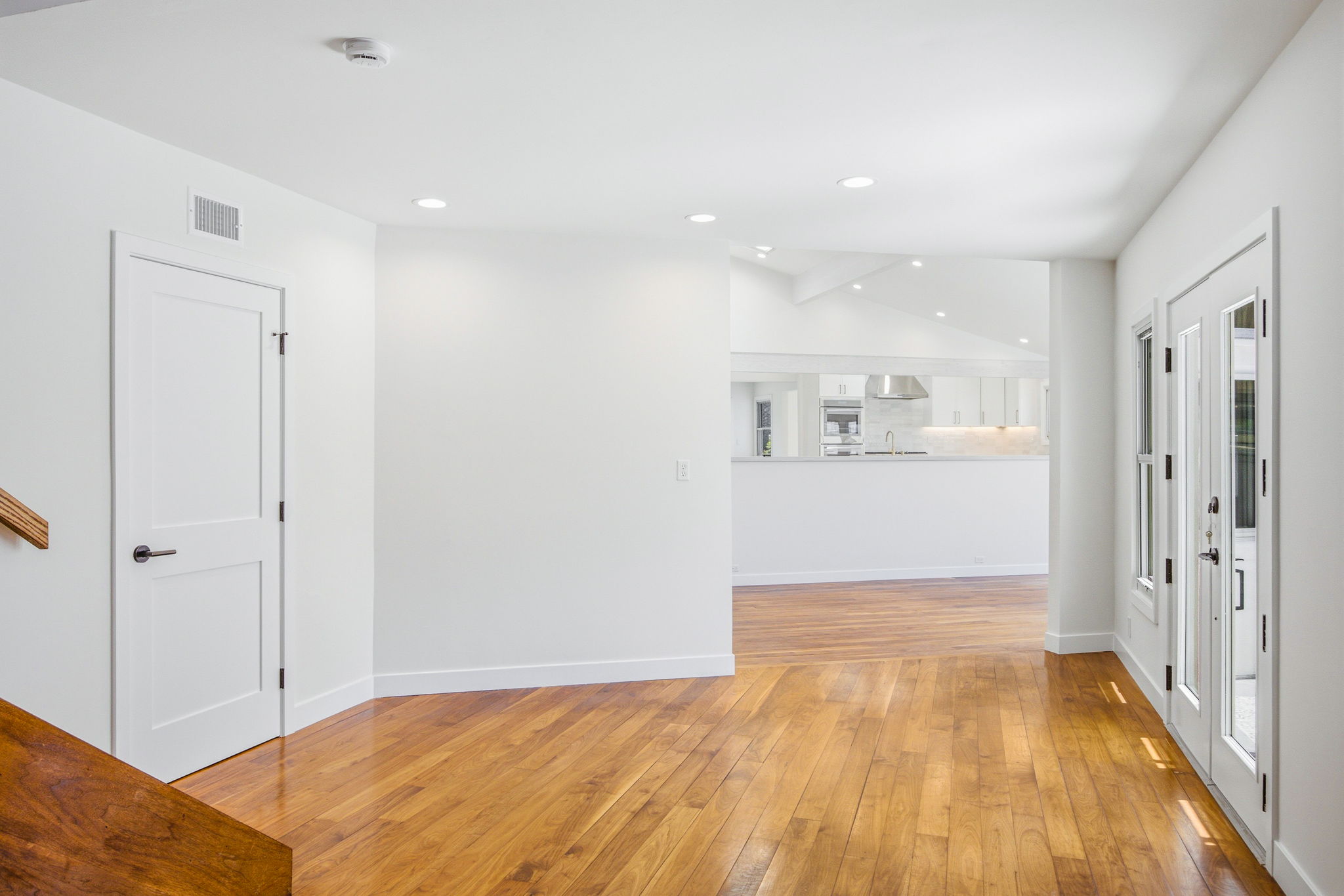 - Oasis Builders, Inc. Bright white hallway with warm wood flooring, doors along the right, and a view into the modern kitchen ahead.