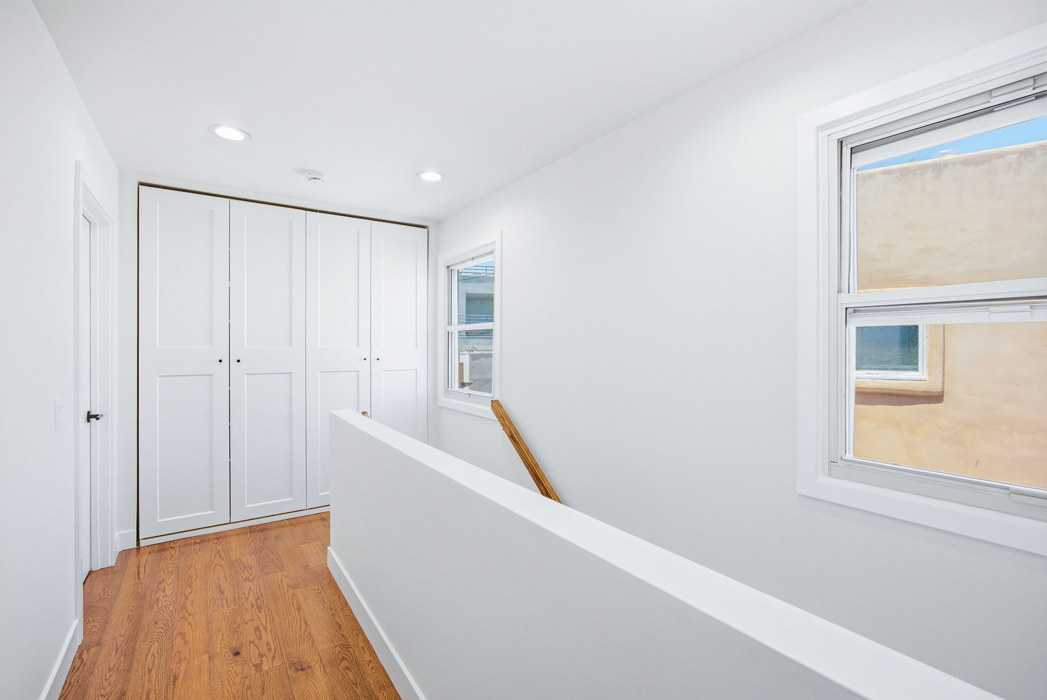 - Oasis Builders, Inc. Bright white hallway with built-in white closet doors on the left, a window on the right, and a wooden floor with a railing along a half-wall.