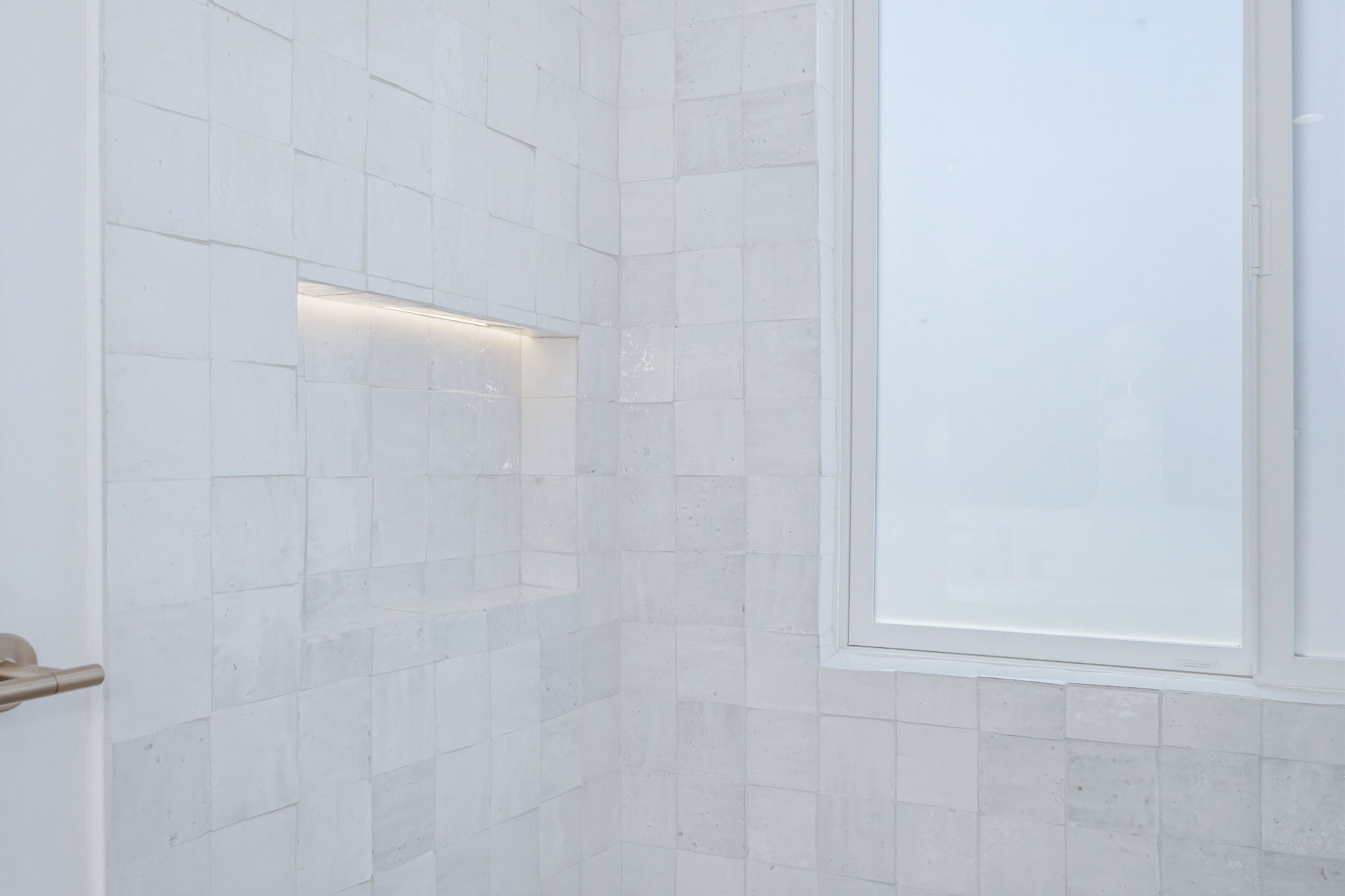 - Oasis Builders, Inc. White marble tiled bathroom wall with a frosted window on the right and a brass door handle visible in the lower left corner; a recessed lit niche runs along the wall.